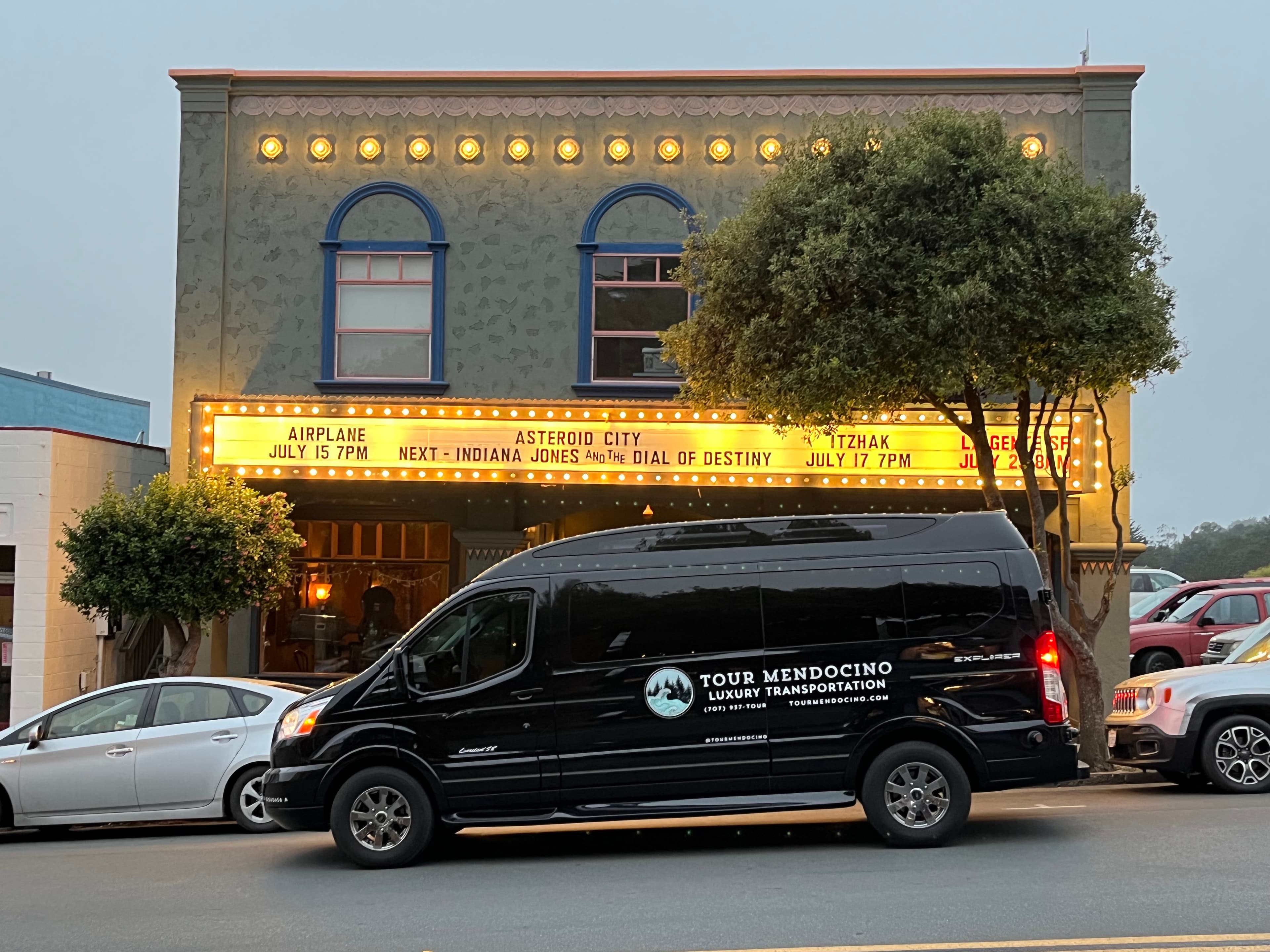 A black van parks in front of a theater displaying upcoming movie titles on its marquee.