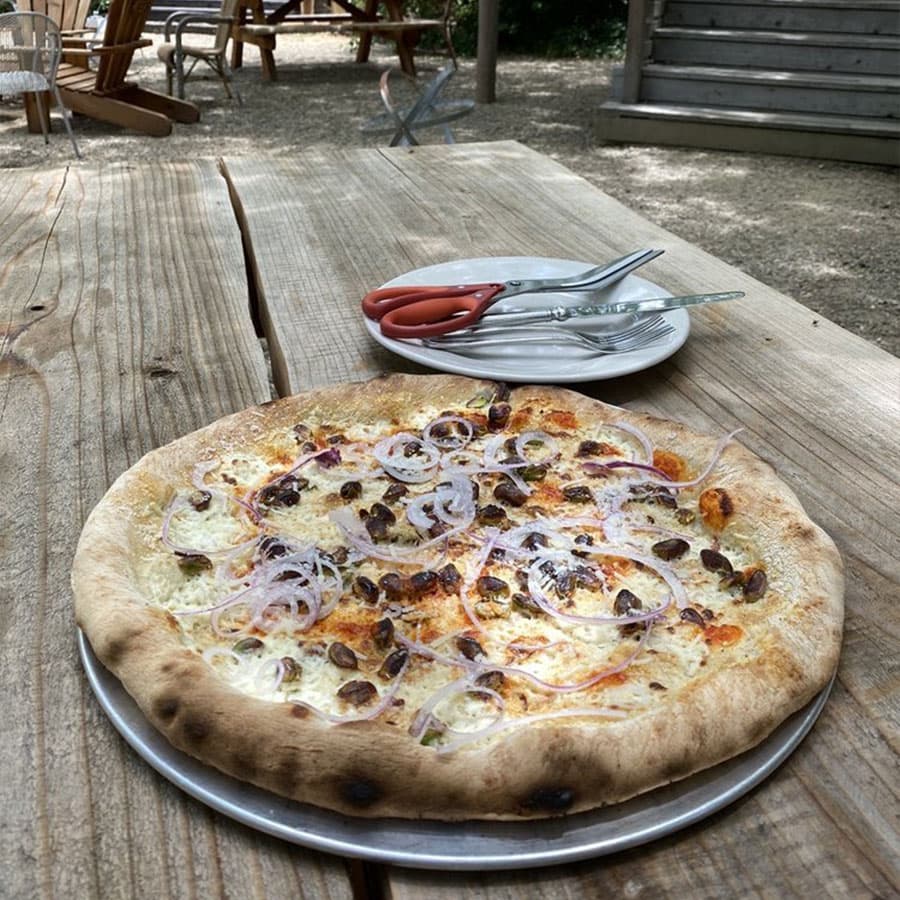 A gourmet pizza with onions, and a white sauce, served on a wooden table at an outdoor restaurant.