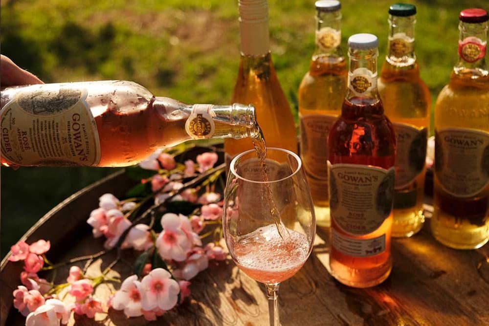 A bottle of Gowans Rose Cider being poured into a glass, with other cider bottles and pink flowers in the background.