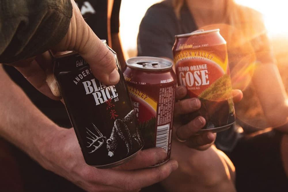 Two people holding cans of Anderson Valley Brewing Company beer: Black Rice Ale and Blood Orange Gose.