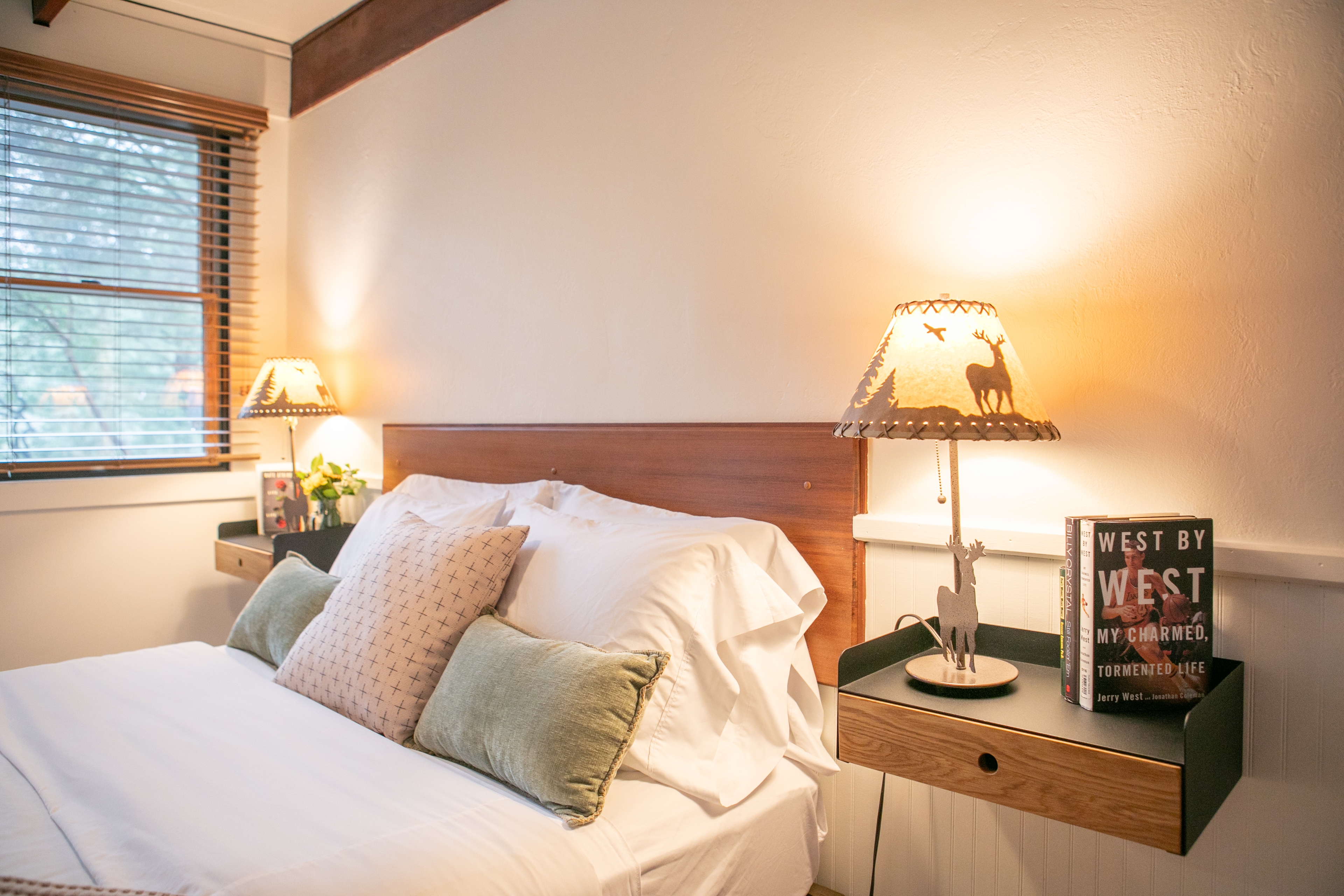 A bed with white sheets and pillows, a wooden headboard, a lamp with a nature-themed shade, and a nightstand with a book.