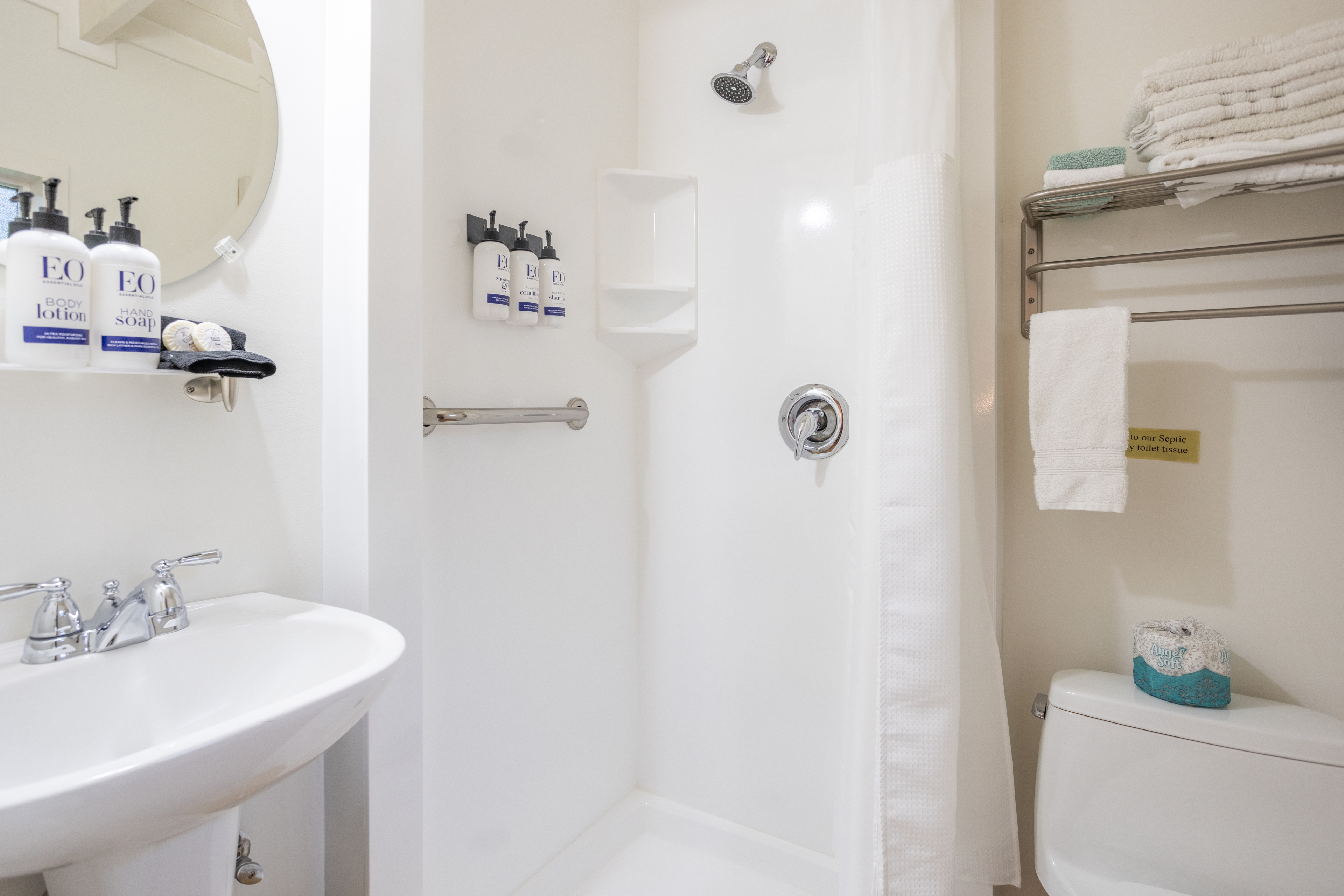 A clean, white bathroom with a shower, toilet, sink, a round mirror, and a shelf with towels.