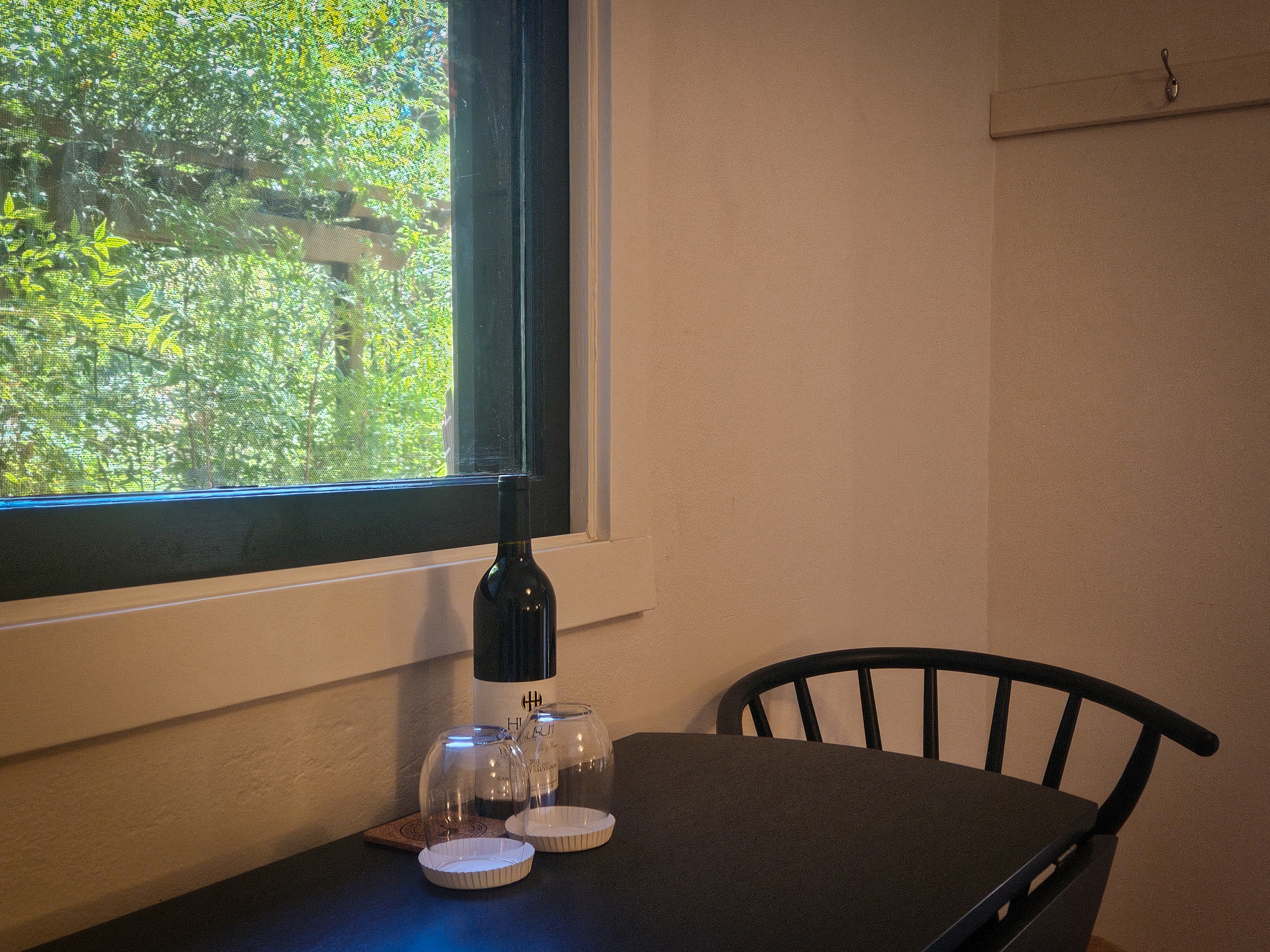 A small table with a bottle of wine, glasses, and a chair next to a window with a view of trees."