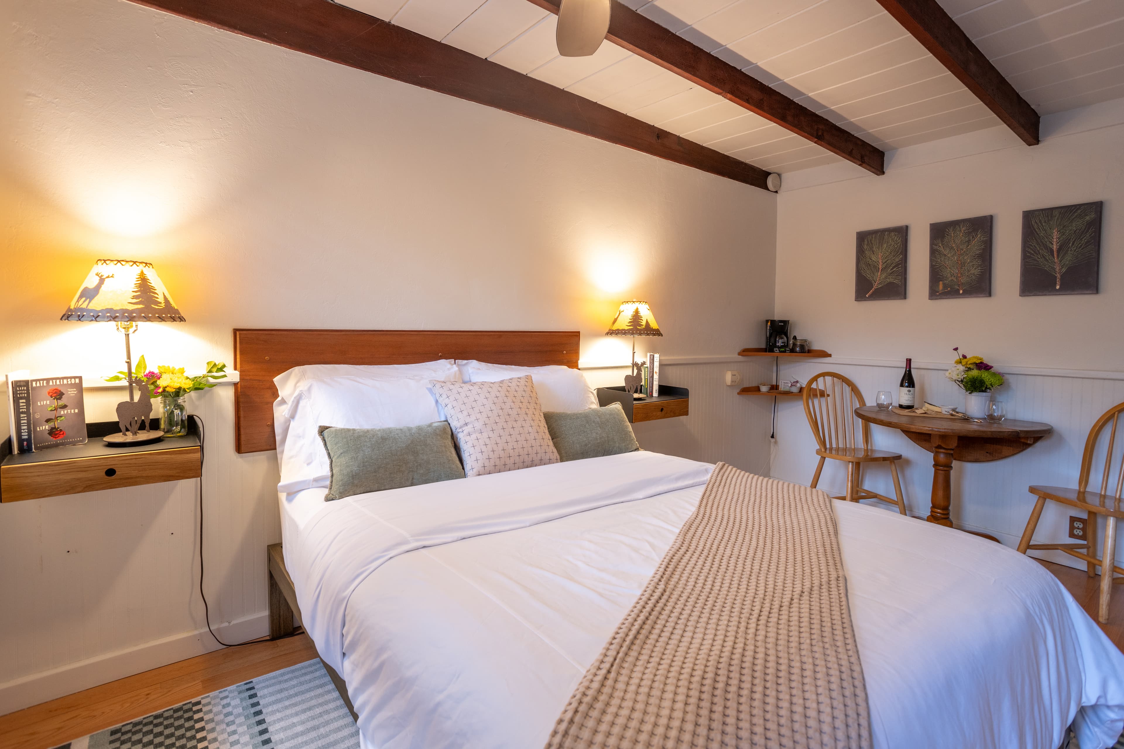 A bedroom with a queen bed with white linens and a tan throw, two nightstands with lamps, a small table with chairs, artwork on the wall, and a wooden beam ceiling.