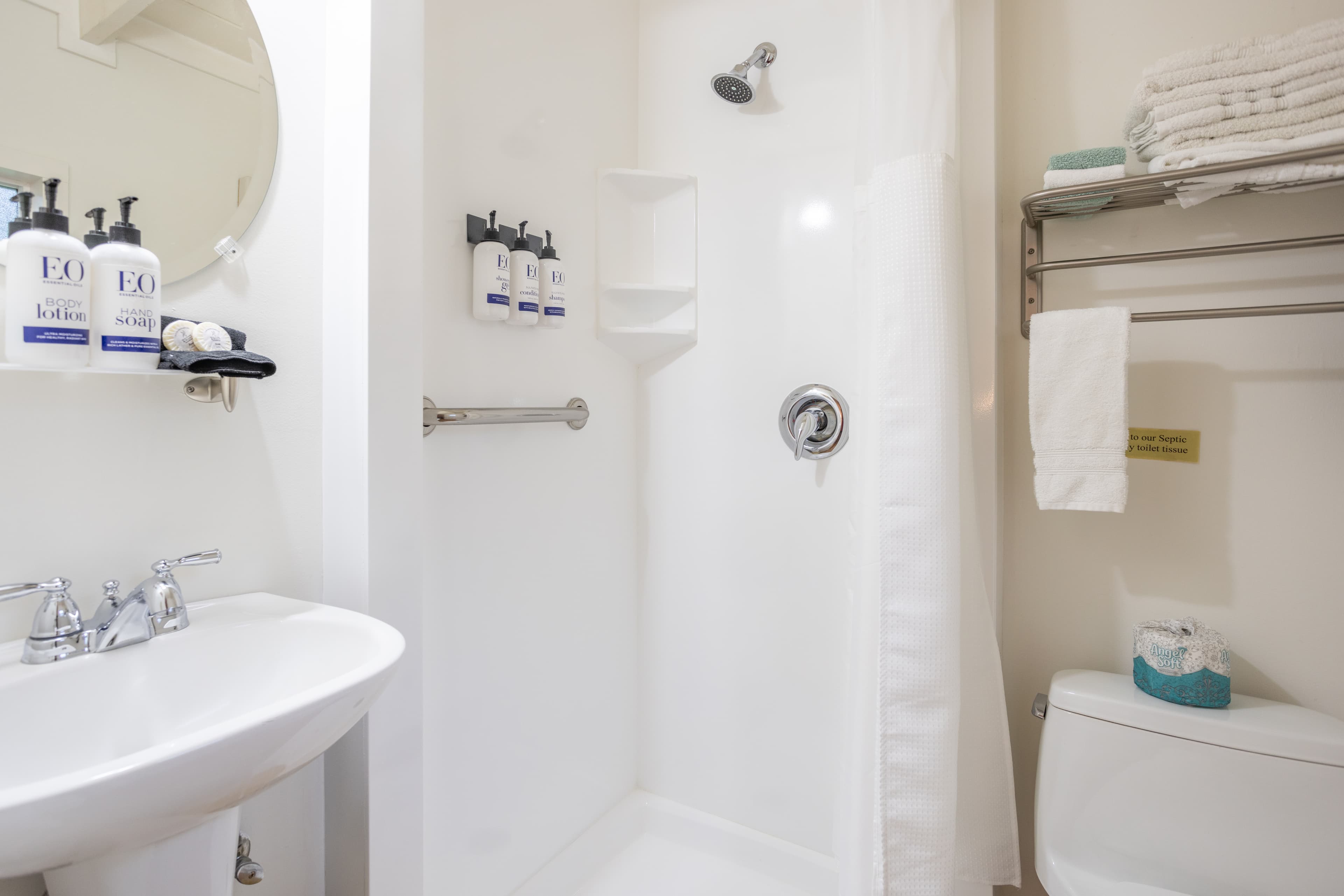 A clean, white bathroom with a shower, toilet, sink, a round mirror, and a shelf with towels.