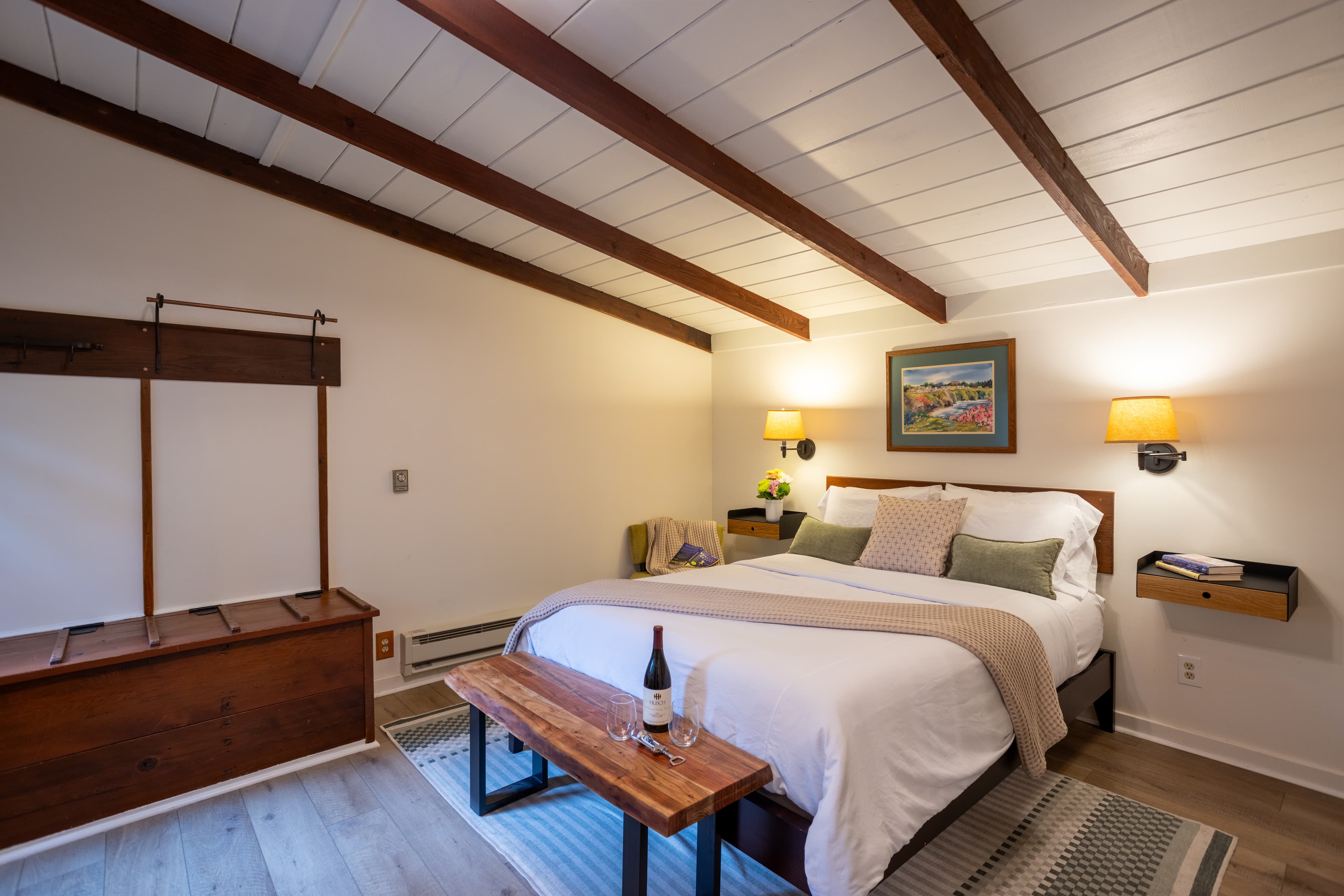 A cozy bedroom with a king-sized bed, wooden furniture, a wine bottle on a bench, and artwork on the wall. The room has a vaulted ceiling with exposed wooden beams.