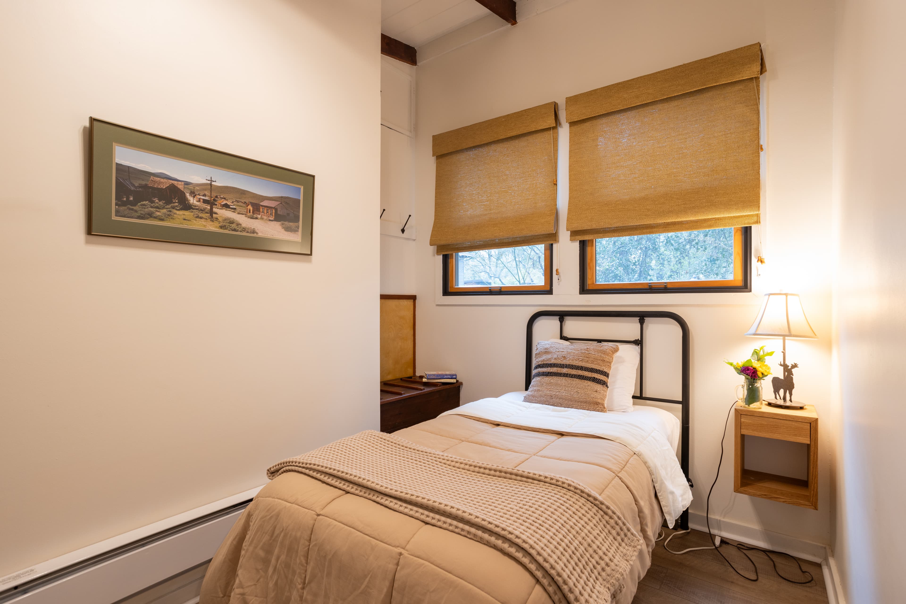 A warm and inviting bedroom with a single bed, wooden furniture, and a window with blinds. The room has a relaxed atmosphere with soft lighting.