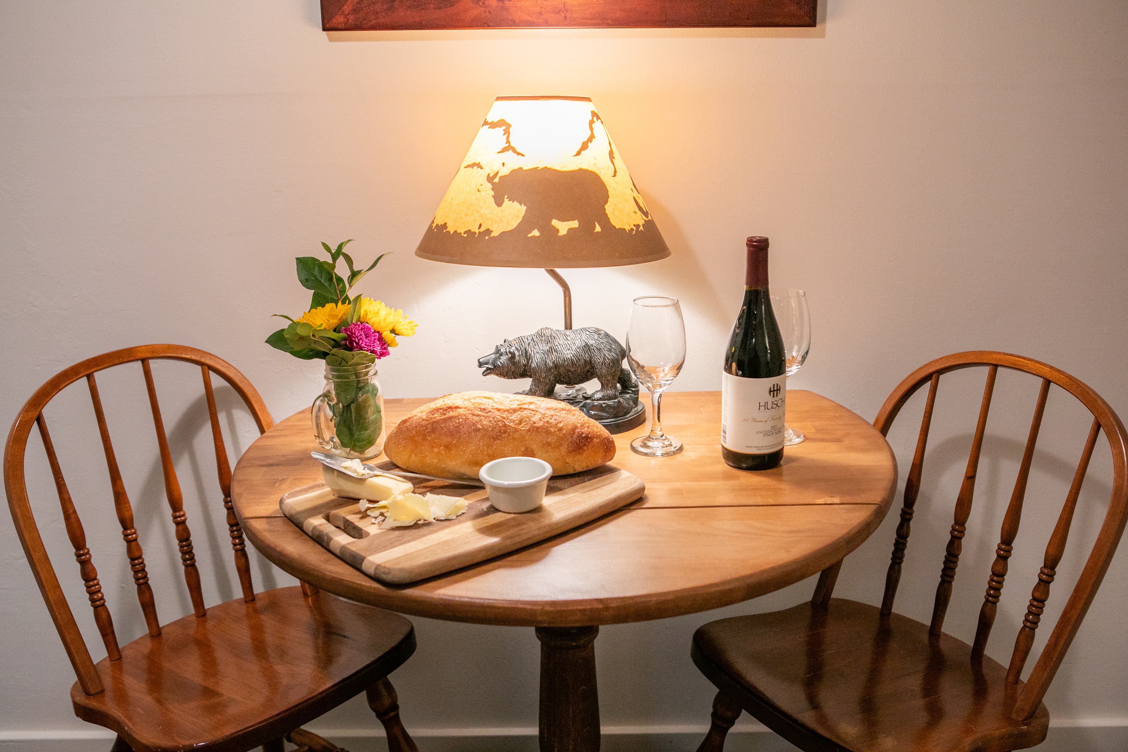 A small table with two chairs, a lamp, a bottle of wine, a loaf of bread, and a vase of flowers.