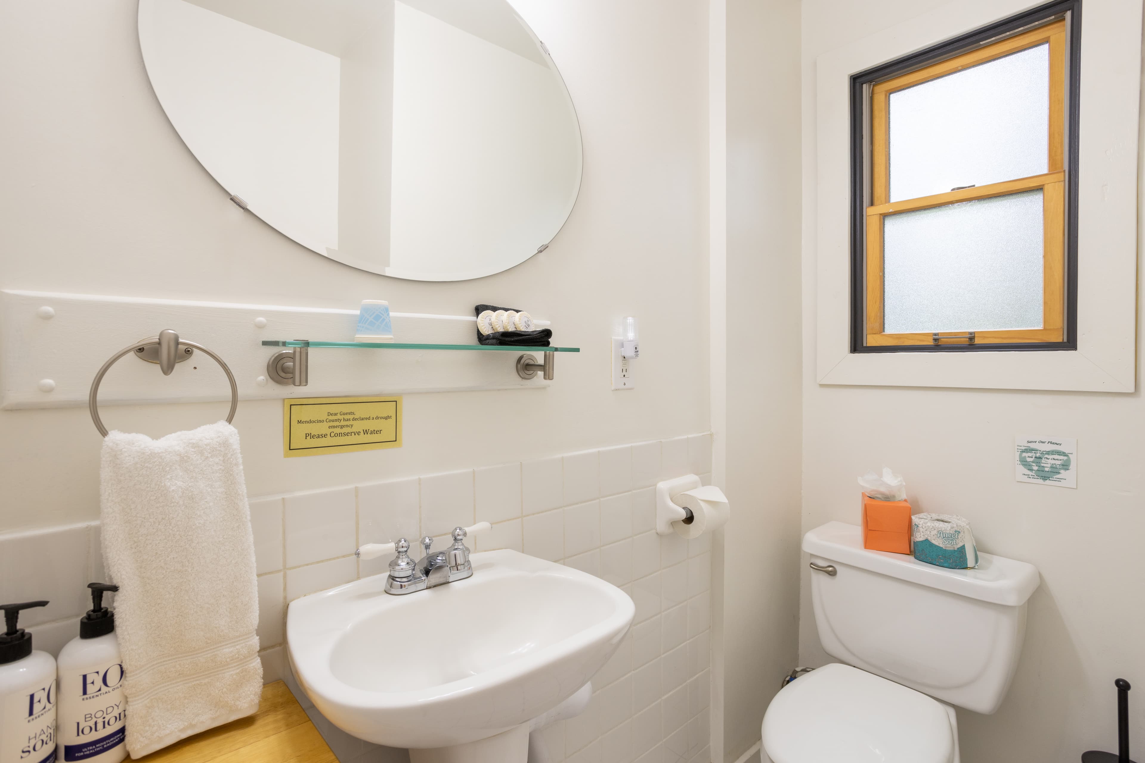 A bathroom with a white sink, a toilet, a large oval mirror, a glass shelf with toiletries, and a window with blinds.