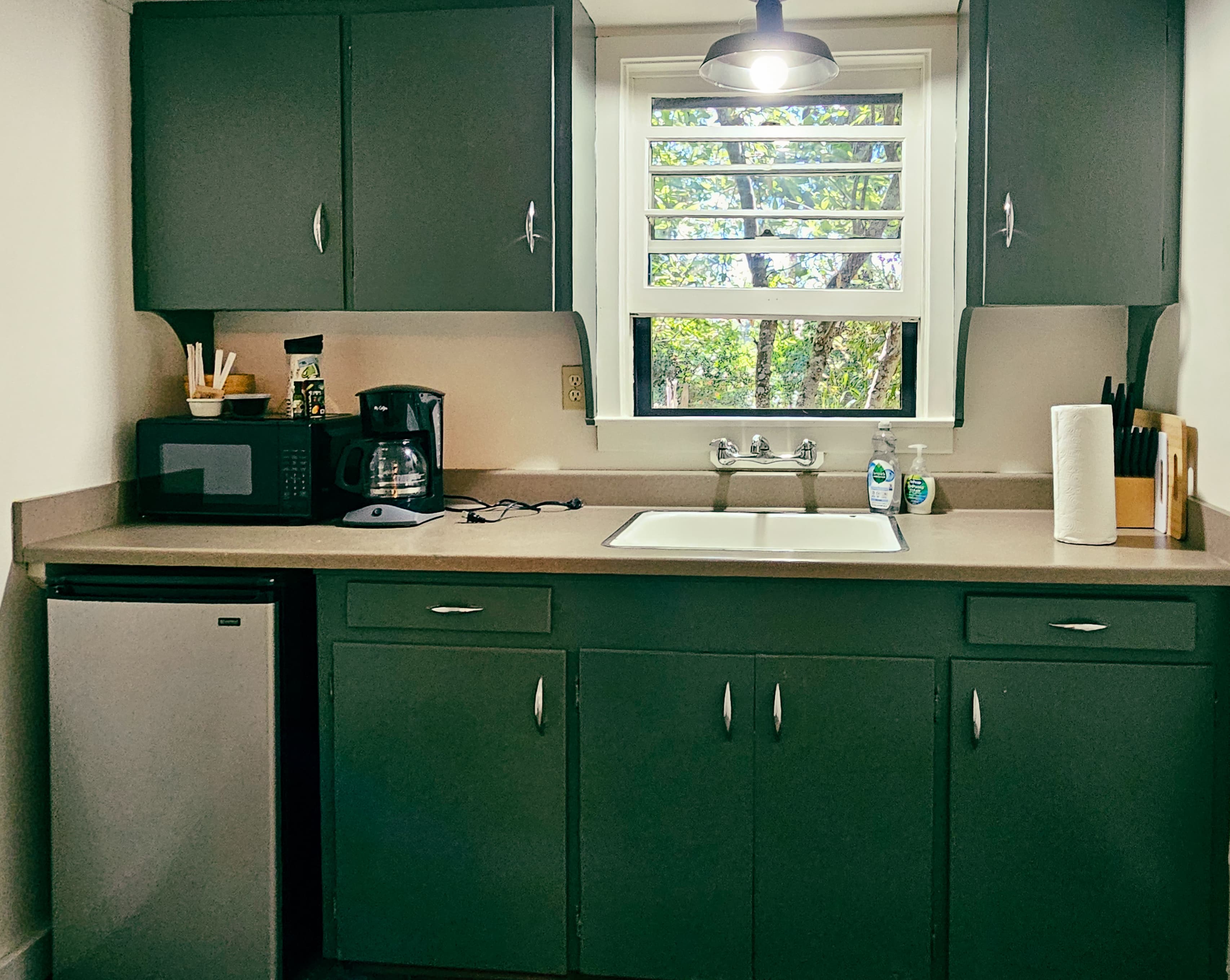 A small kitchen with dark green cabinets, a sink, a microwave, a coffee maker, a mini-fridge, and a window with shutters.