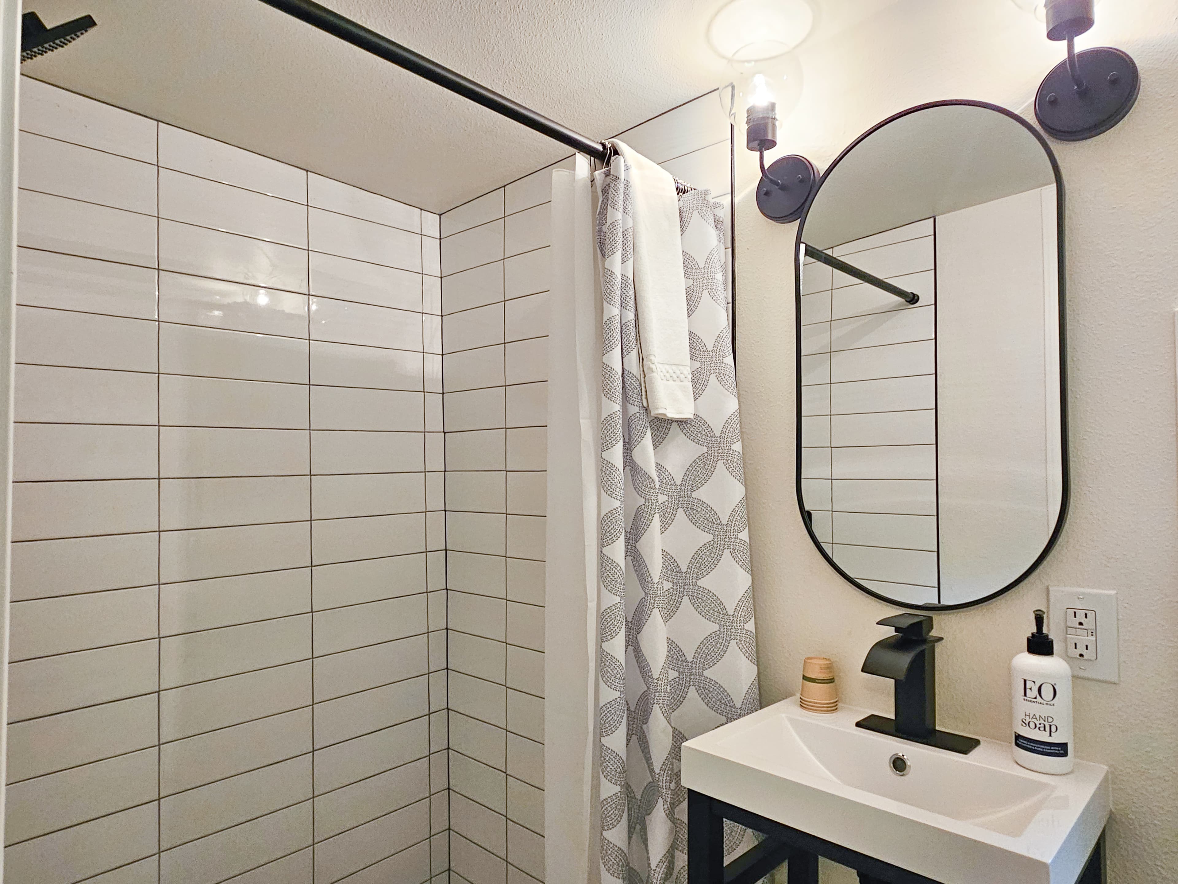 A bathroom with a tiled shower, a sink, and an oval mirror with black sconces on either side.