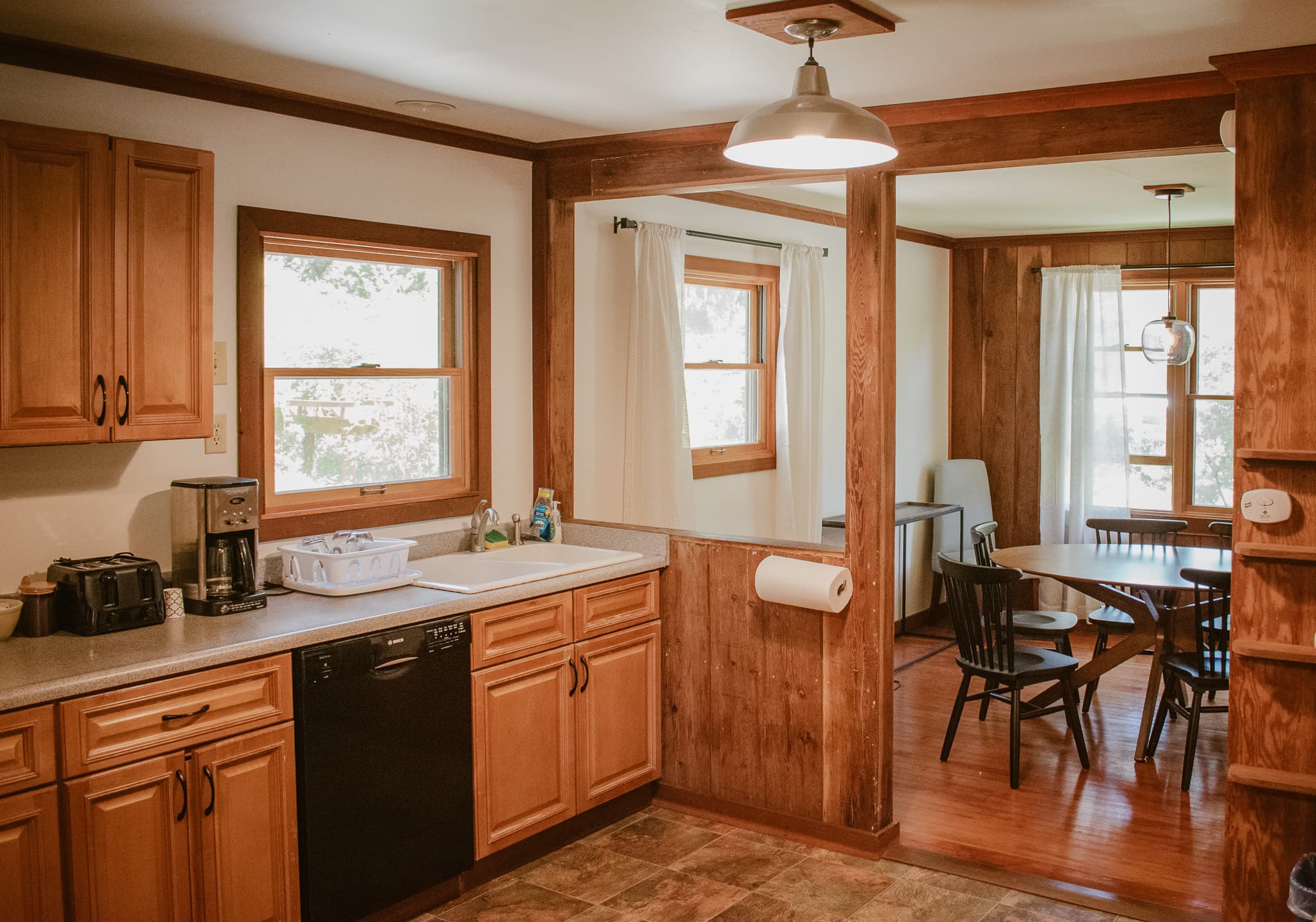 A kitchen with wooden cabinets, a sink, a dishwasher, and a coffee maker, opening into a dining area with a table and chairs.