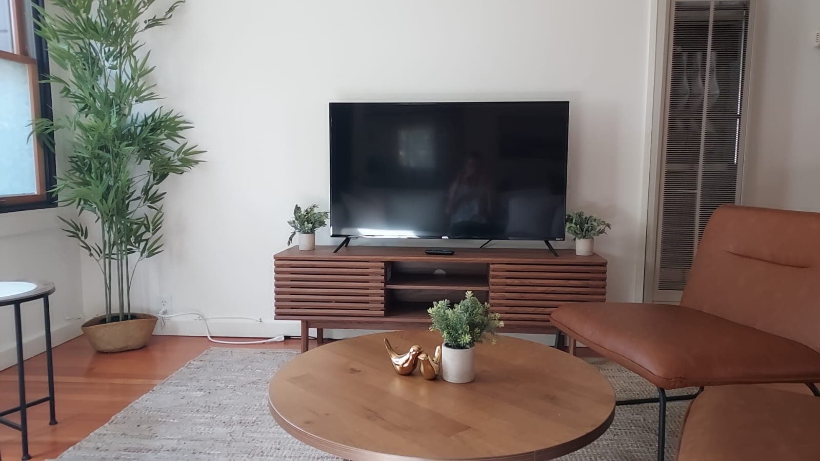 A living room with a large flat-screen TV on a wooden stand, a brown leather armchair, a round coffee table with decor, and a tall plant in a corner.