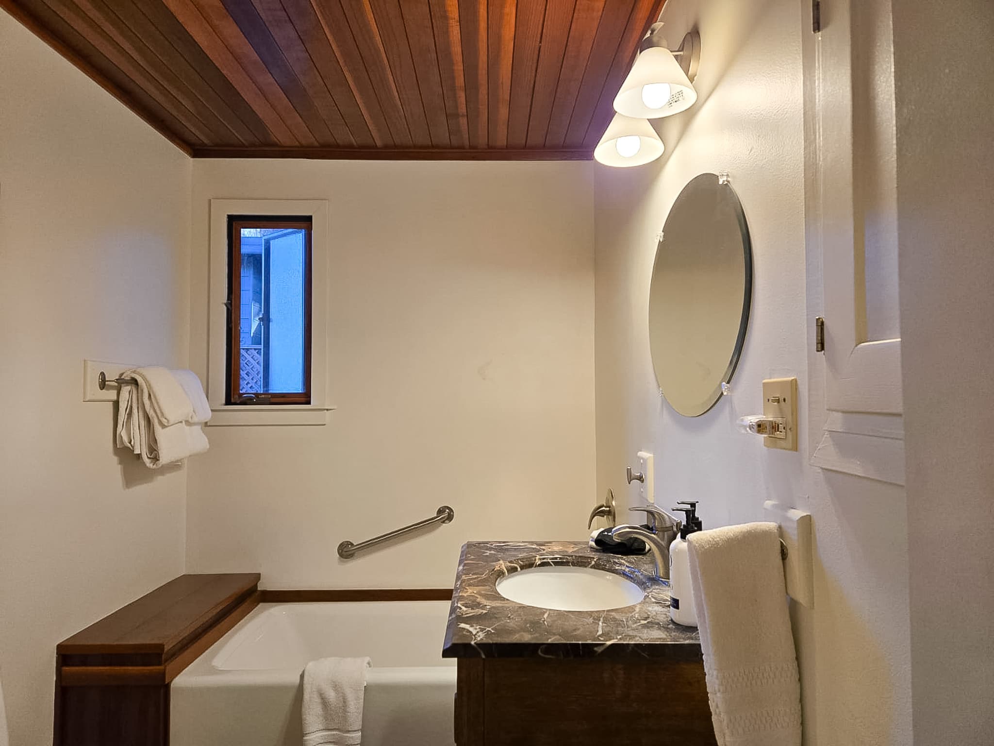 A bathroom with a bathtub, a sink with a marble countertop, a round mirror, a window, and a grab bar next to the bathtub.