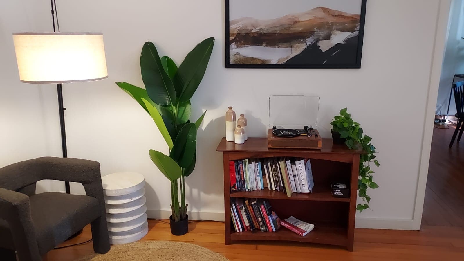 A living room corner with a floor lamp, a tall leafy plant, a wooden bookshelf with books and decor, and a small plant on top of the shelf.