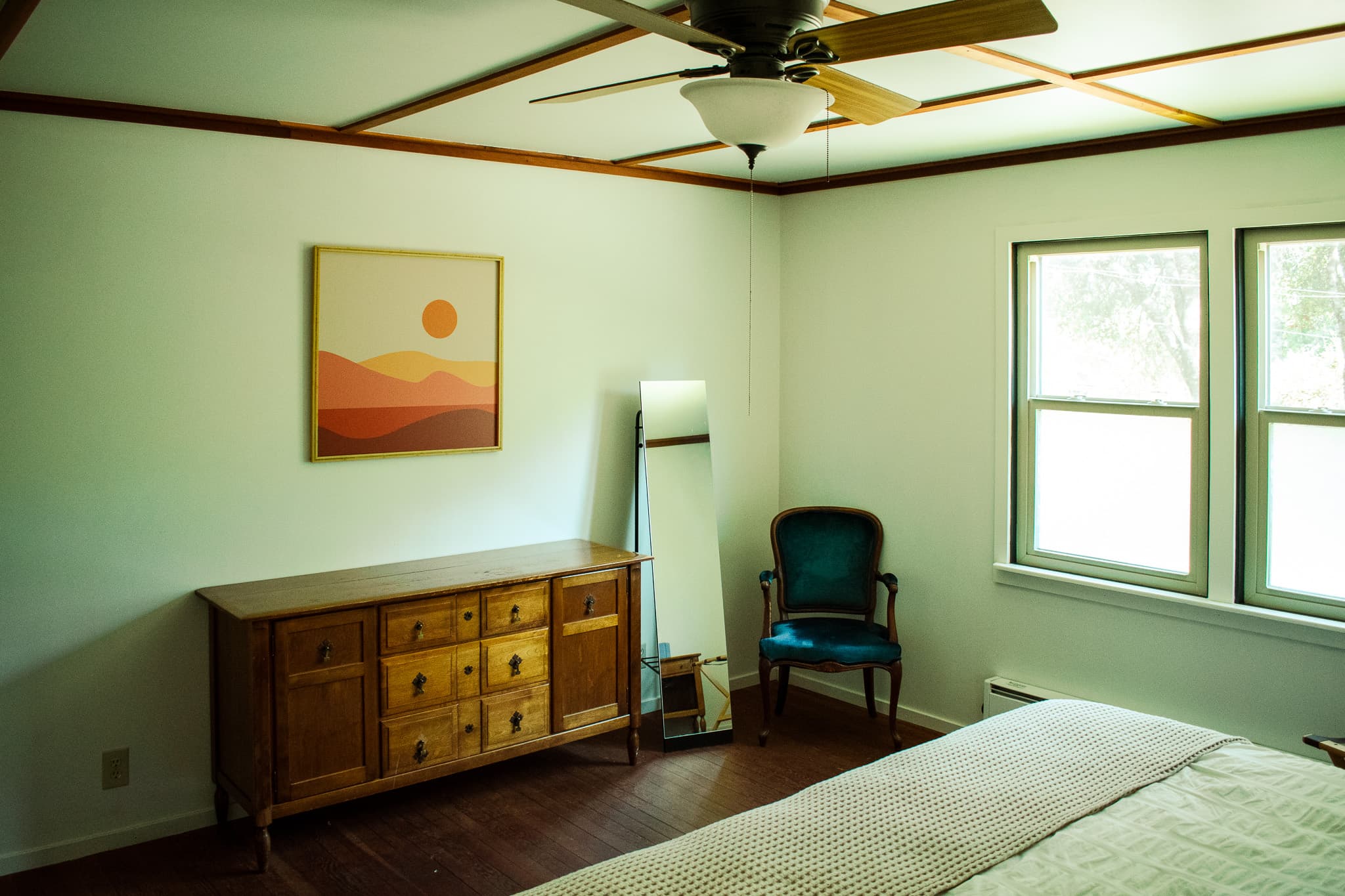 A bedroom with a bed, a wooden dresser, a blue armchair, a full-length mirror, a painting of a sunset, and a ceiling fan.