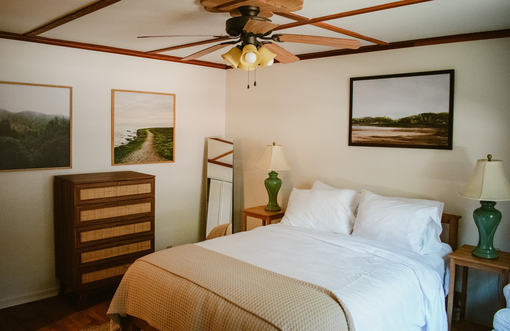 A bedroom with a queen bed with white bedding and a beige throw, two nightstands with lamps, a dresser, artwork depicting landscapes, and a ceiling fan.