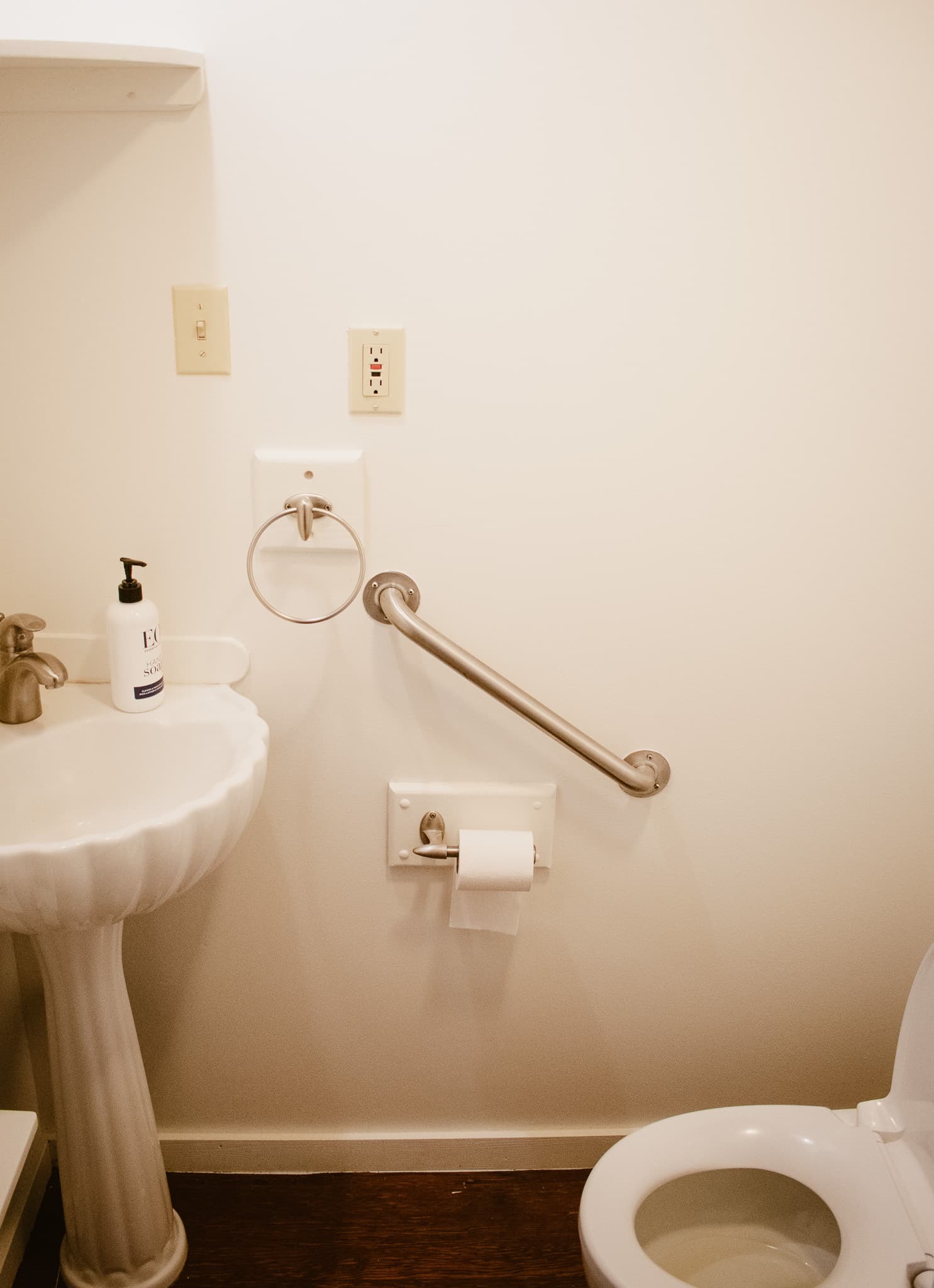 A bathroom with a white pedestal sink, a toilet, a grab bar on the wall, and a towel rack.