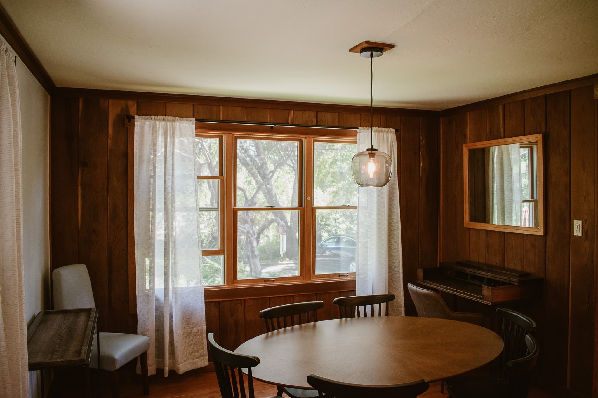 A dining room with a round wooden table and chairs, a large window with white curtains, and wood paneling on the walls.