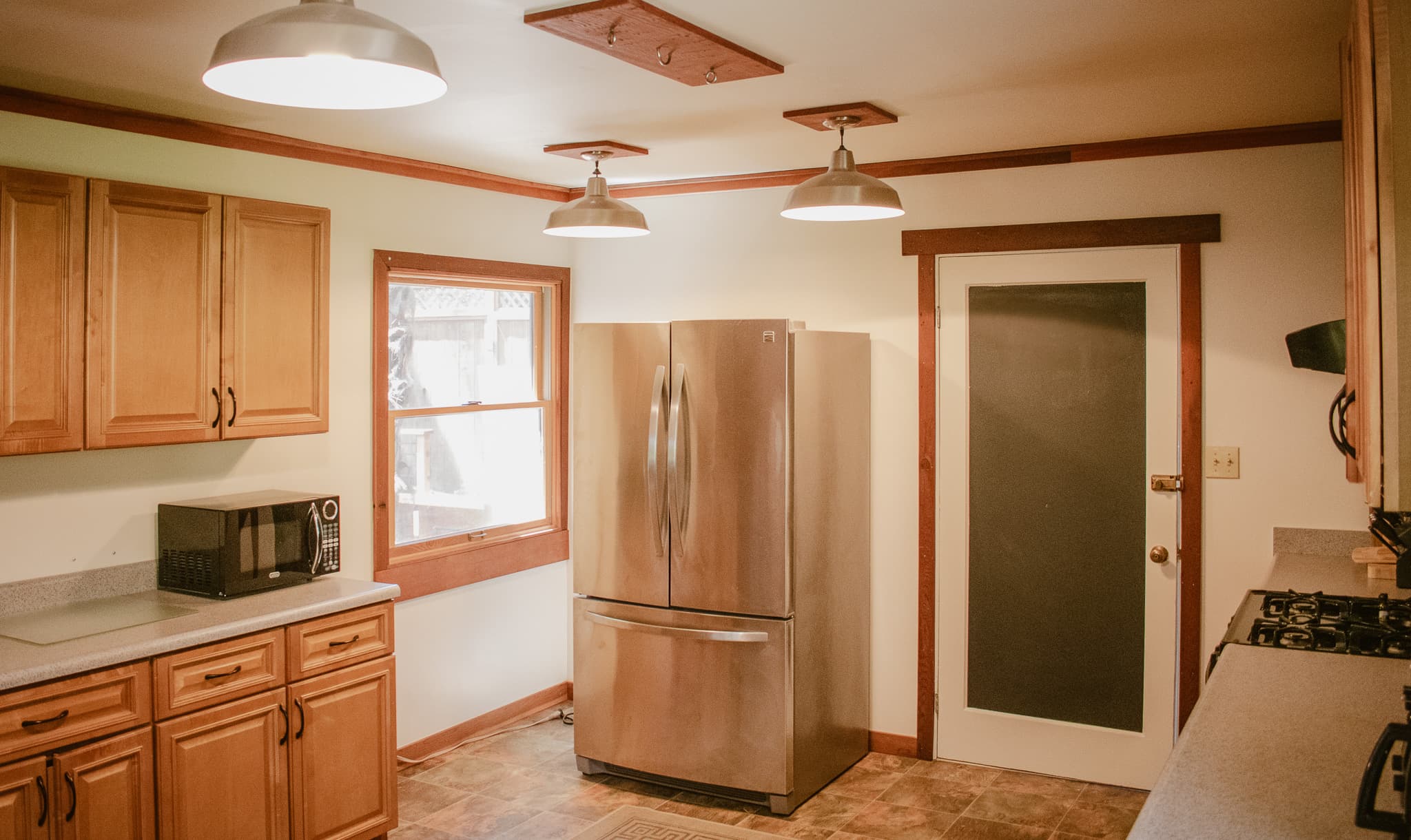 A kitchen with wooden cabinets, a stainless steel refrigerator, a microwave, a stove, and a window with a view of trees outside.